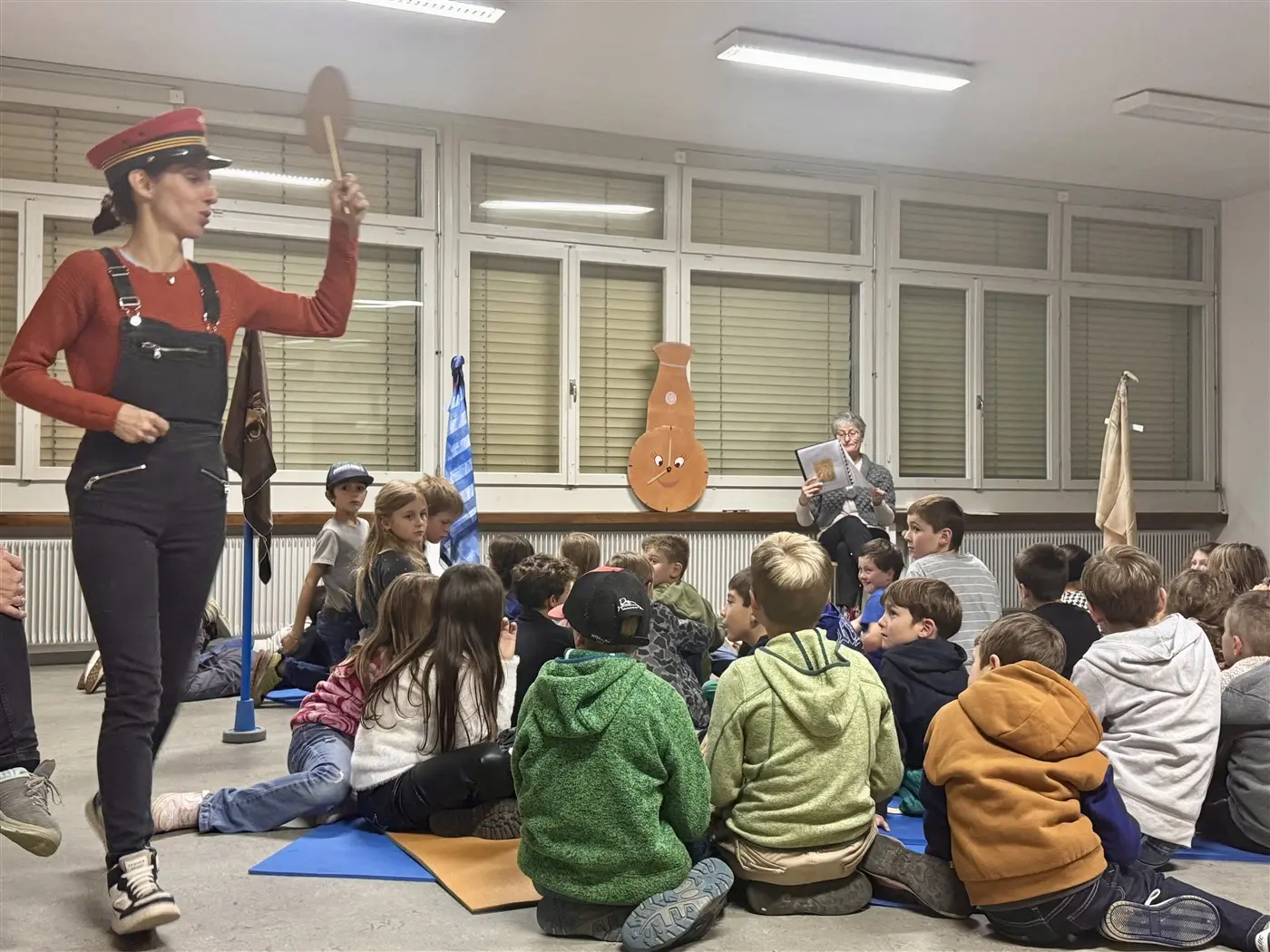Una persona in costume da ufficiale, con un cappello e una paletta, sta dirigendo un gruppo di bambini seduti a terra. Sullo sfondo, si vede una donna che legge un libro, con decorazioni murali sul tema. I bambini seguono attentamente l'attività