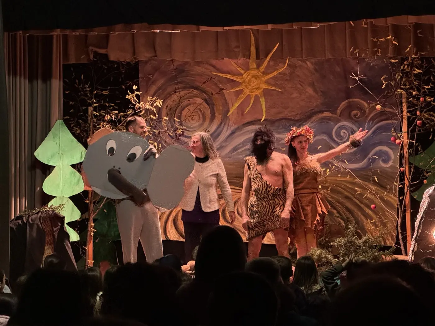 Un palco teatrale con decorazioni naturali. Un uomo vestito da elefante interagisce con tre performance: una donna con abiti preistorici e due personaggi, uno con una maschera nera e l'altro in abiti di foglie. Il pubblico è