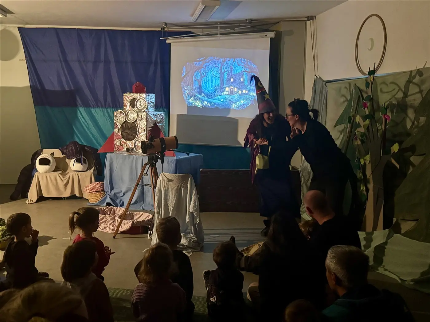 Un gruppo di bambini seduti a terra guarda uno spettacolo teatrale. Sul palco, due attrici, una con un cappello da strega, interagiscono mentre una scenografia incantevole viene proiettata sullo sfondo.