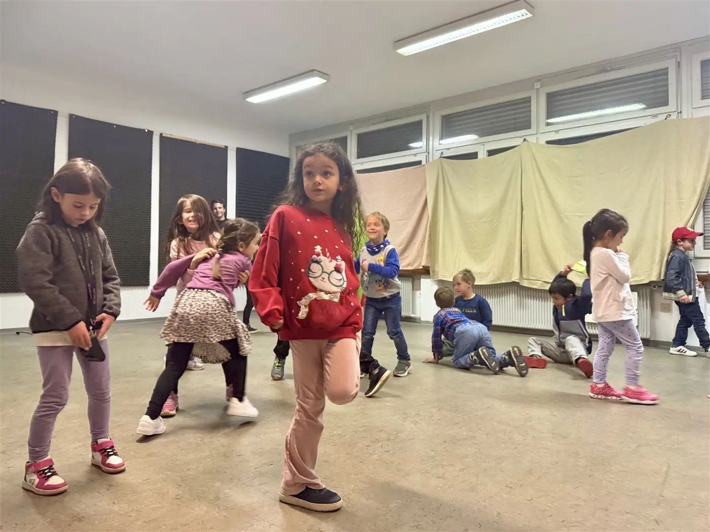 Un gruppo di bambini sta ballando in una stanza. Alcuni sono in movimento, mentre altri sembrano osservare. Alcuni indossano vestiti colorati e ci sono diverse espressioni di concentrazione e divertimento.