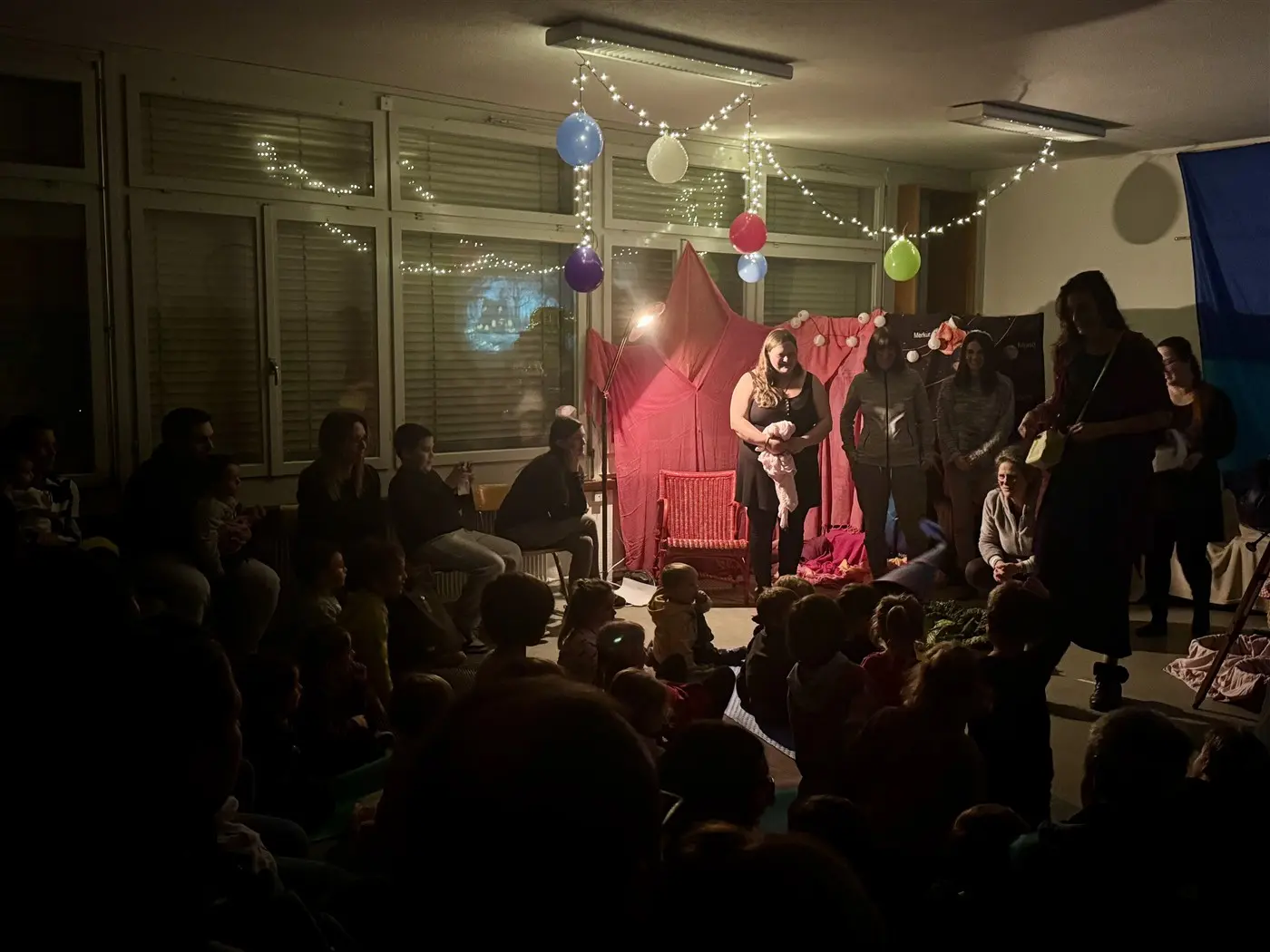 Uno spazio interno con un gruppo di persone sedute, mentre alcune si esibiscono su un palco con decorazioni colorate. La scena è illuminata da luci e palloni appesi, creando un'atmosfera festosa. I bambini in primo piano osservano attentamente.