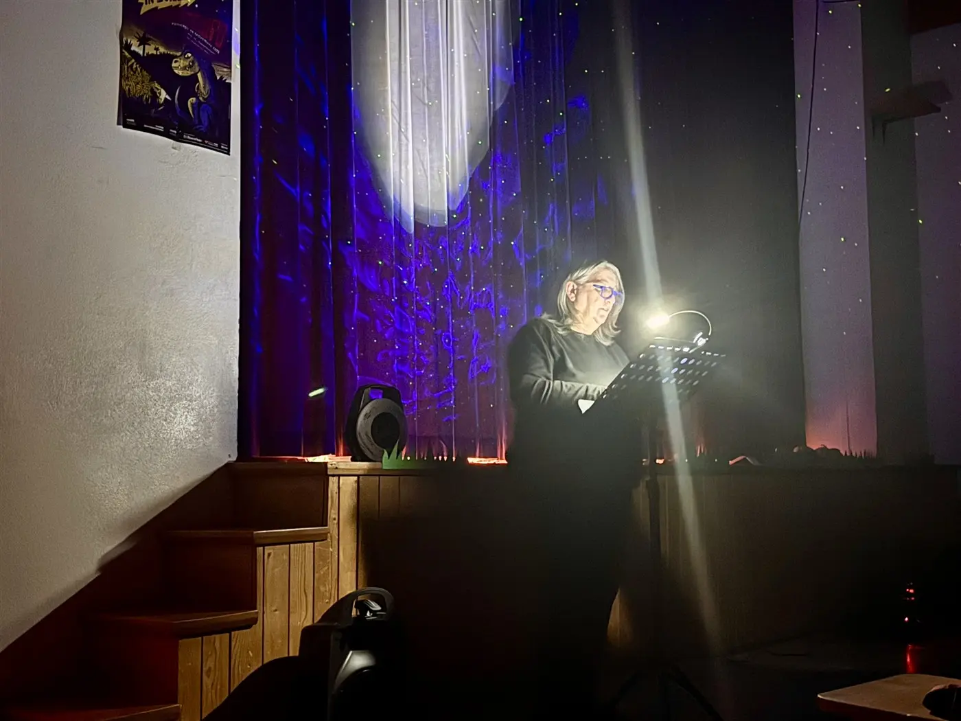 Una persona legge appoggiata a un leggio, illuminata dalla luce di uno spot, in un ambiente con tende scure e decorazioni luminose. Sullo sfondo si vedono gradini in legno e un altoparlante.