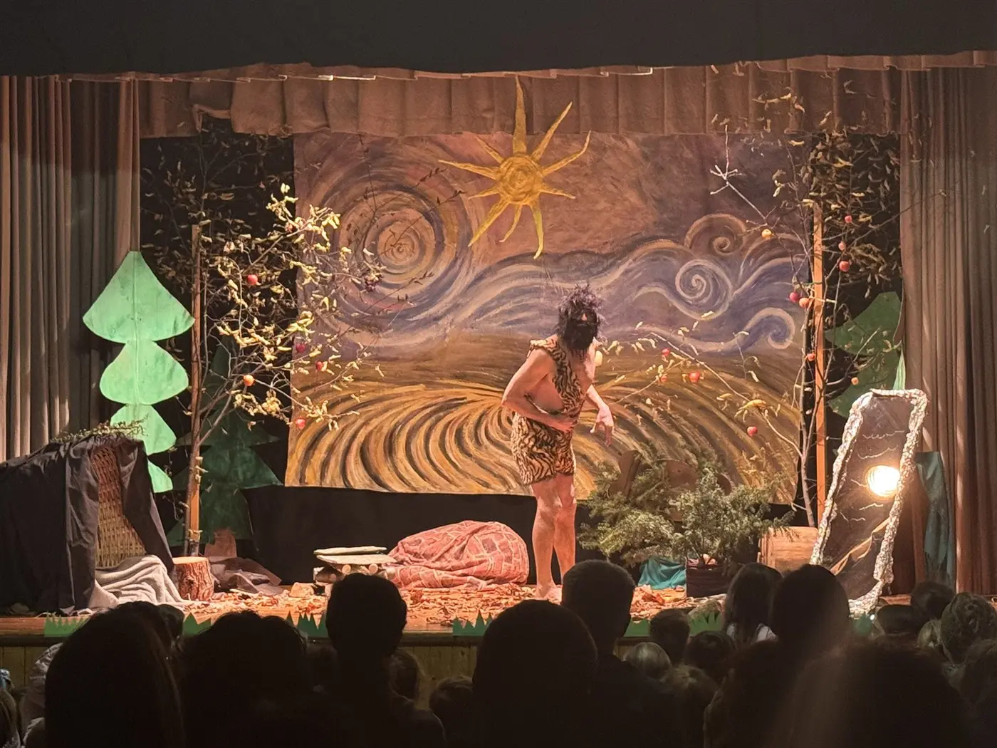 Una scena teatrale con un'attrice in costume che si esibisce su un palcoscenico decorato con elementi naturali, come alberi di cartone e un grande sfondo dipinto raffigurante un paesaggio. Il pubblico osserva attentamente.