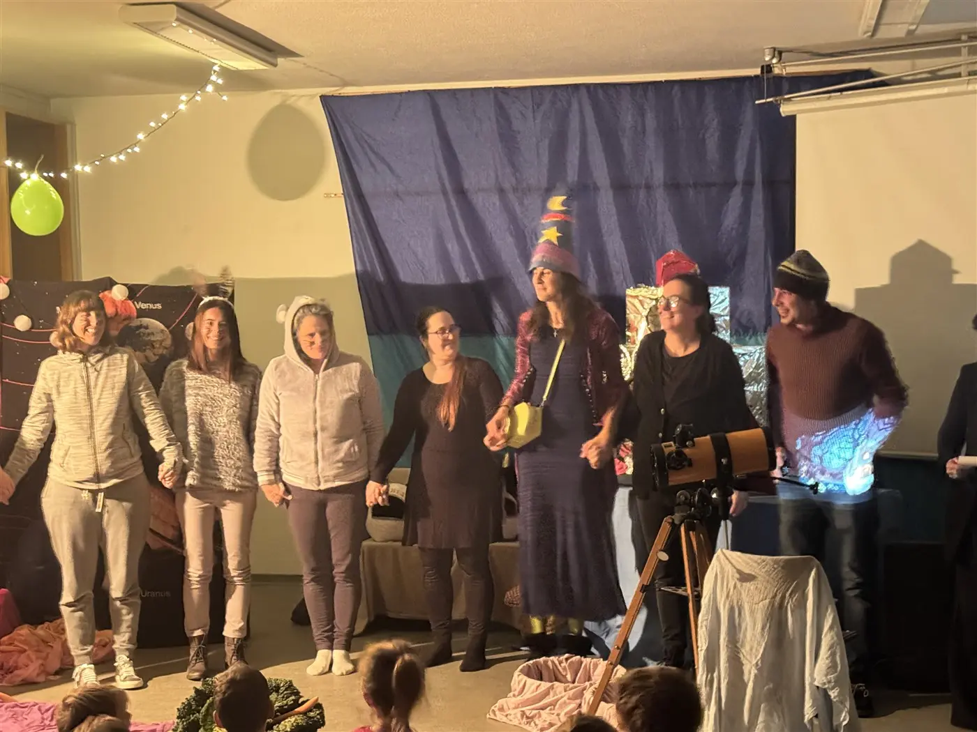 Un gruppo di persone in abiti festivi si tiene per mano sul palco, sorridendo. Diverse decorazioni colorate e un telescopio sono visibili sullo sfondo. I bambini si trovano seduti in primo piano, osservando attentamente.