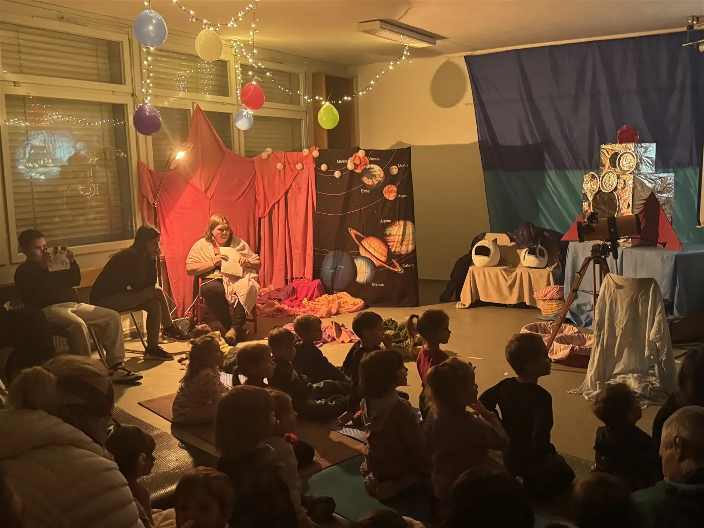 Un gruppo di bambini seduti a terra guarda affascinato uno spettacolo. Alcuni adulti, tra cui una donna che tiene un libro, sono seduti su sedie mentre lo sfondo è decorato con tende rosse e illustrazioni di pianeti. Luci soff