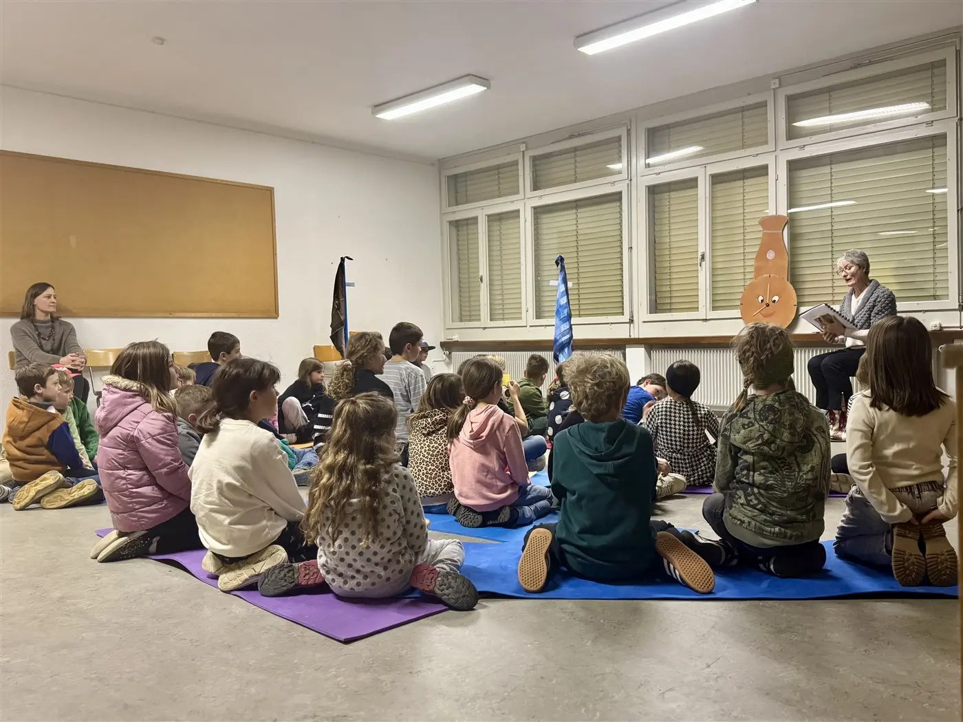 Un gruppo di bambini ascolta attentamente un racconto da un narratore seduto su una sedia. Alcuni adulti li osservano. La stanza è luminosa e decorata con oggetti colorati.