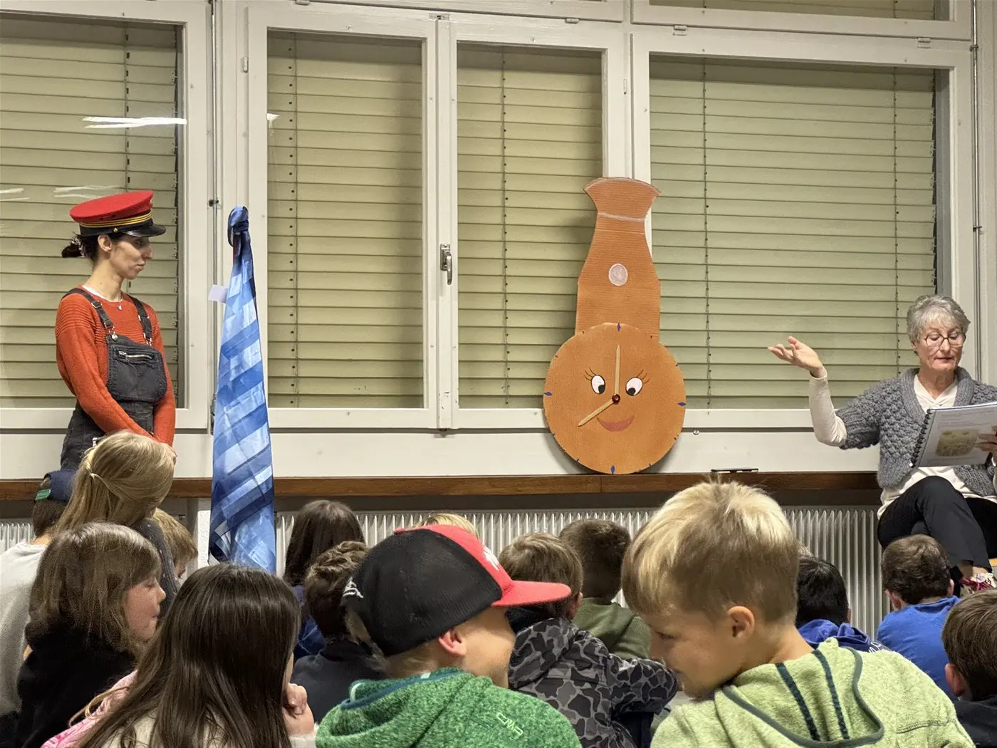 Una narratrice legge una storia a un gruppo di bambini seduti. A sinistra, una donna in un cappello rosso e tuta osserva con interesse. Sul muro è appesa una grande figura di un pesce con un volto sorridente.