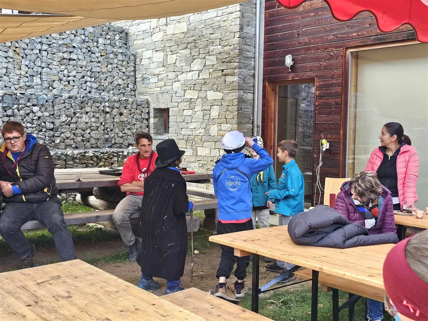 Gruppo di persone in un'area all'aperto, alcune sedute a un tavolo di legno e altre in piedi. Due bambini indossano un cappello e una giacca blu, mentre un adulto indossa una maglietta rossa. Lo sfondo presenta una parete di pietra e una costruzione in legno.