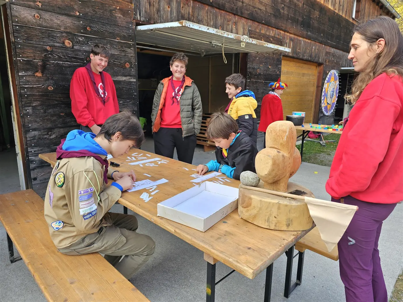 Gruppo di ragazzi che partecipano a un'attività all'aperto, seduti a un tavolo di legno. Alcuni disegnano, mentre altri osservano. Gli adulti sorridono e interagiscono con i ragazzi. Sullo sfondo, una struttura di legno e alcuni oggetti colorati.