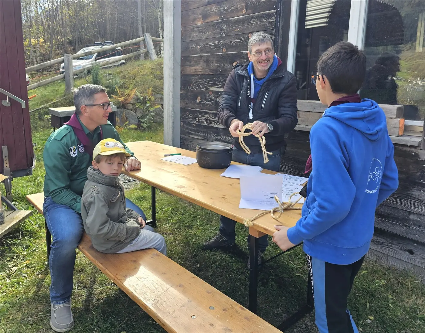 Un uomo sorridente mostra delle corde a un bambino e a un ragazzo davanti a un tavolo all'aperto. Due adulti osservano con interesse. Sullo sfondo, ci sono alberi e una costruzione in legno.