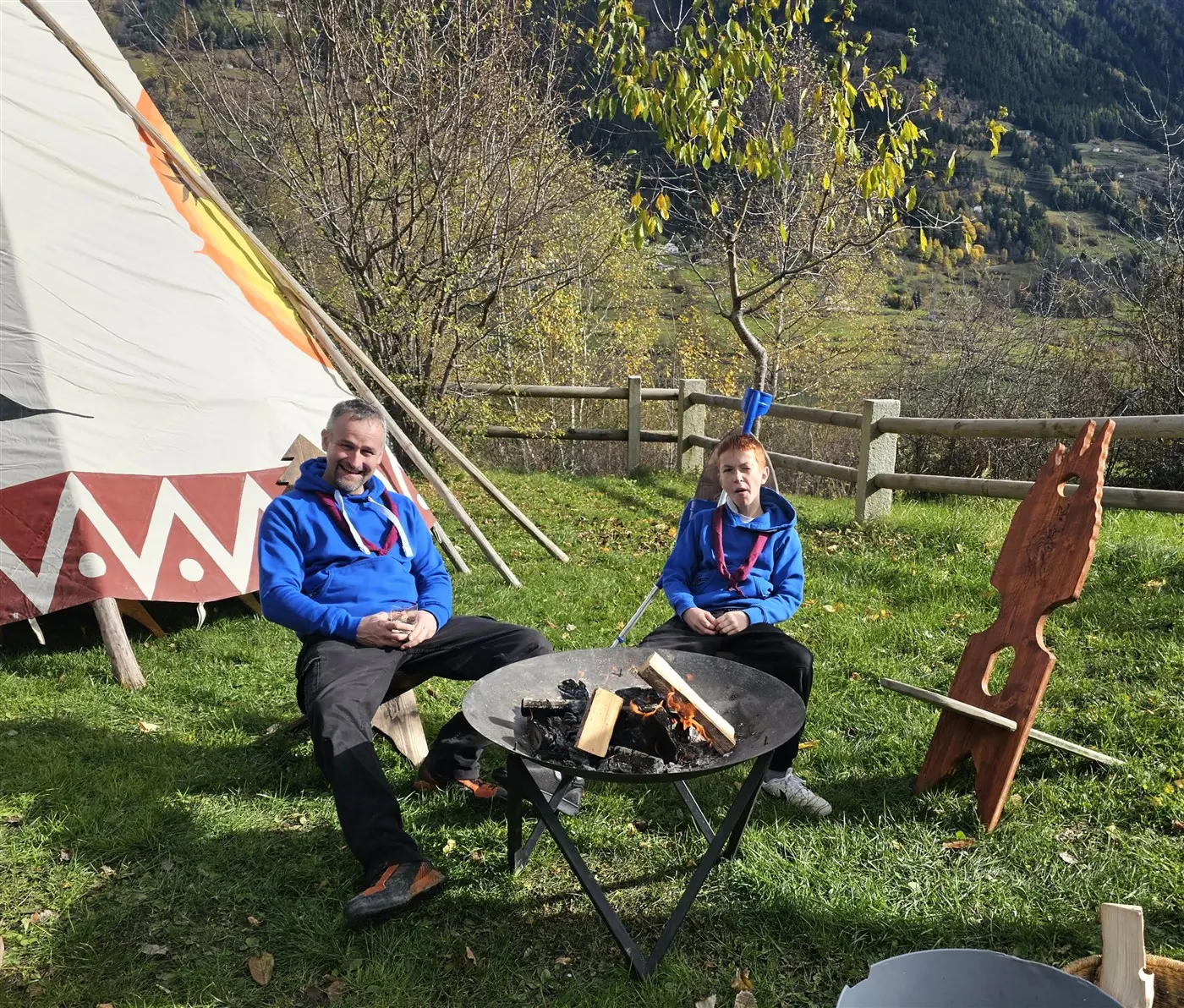 Due uomini seduti attorno a un fuoco, sorridenti. Uno di loro è un adulto e l'altro un bambino, entrambi indossano giacche blu. Sullo sfondo si vede un teepee e alberi verdi.
