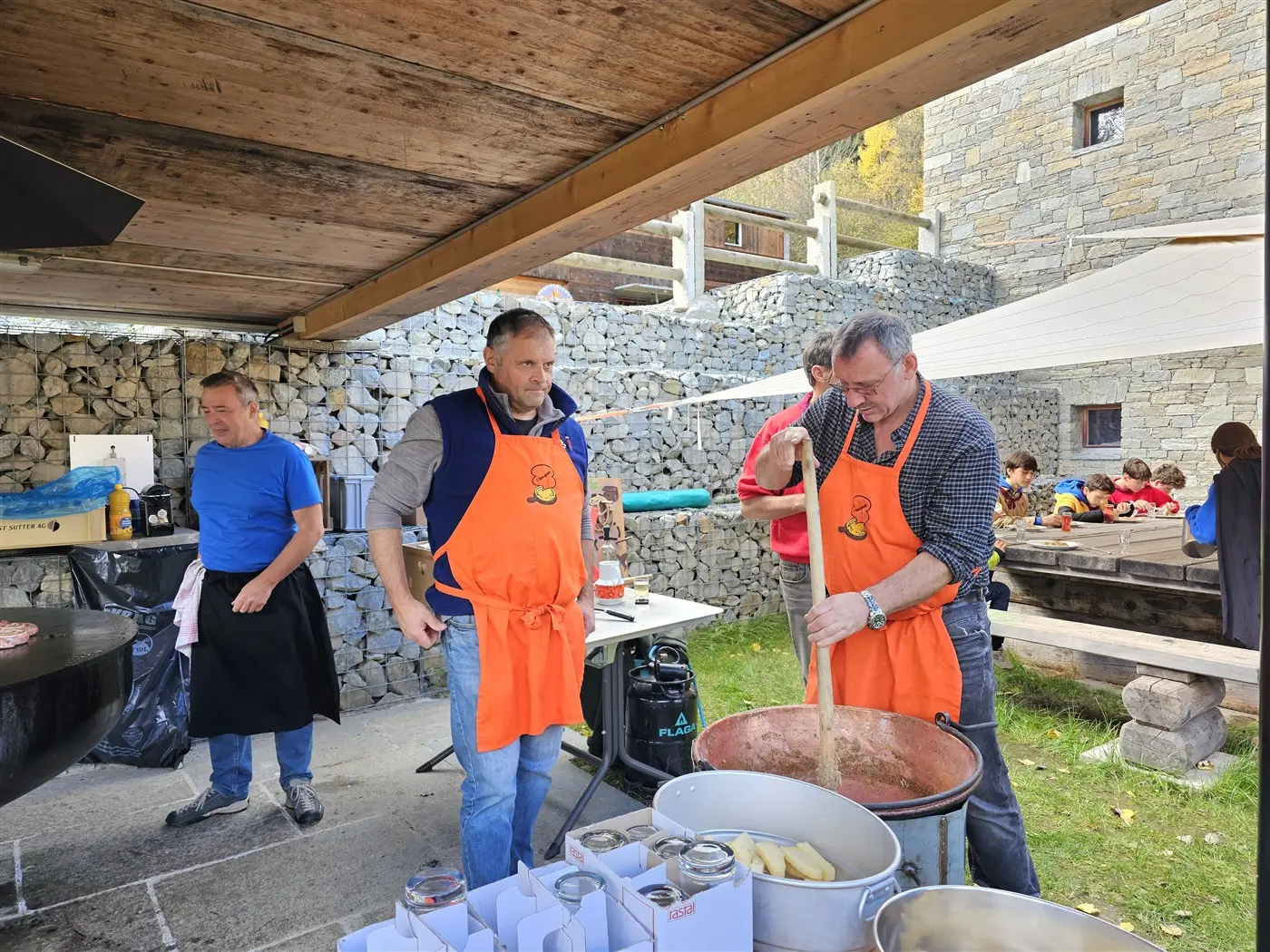 Uomini in grembiuli arancioni cucinano all'aperto, mescolando ingredienti in una grande pentola. Sullo sfondo, un gruppo di persone è seduto a tavoli di legno. La scena si svolge in un ambiente rurale con pareti in pietra.