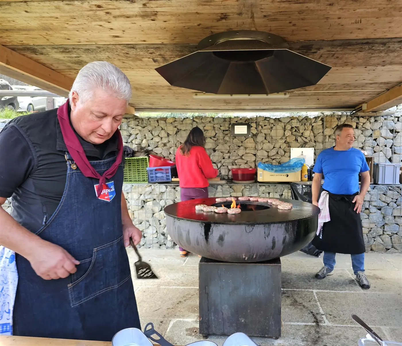 Un cuoco prepara cibo su una griglia rotonda in un'area all'aperto. Due uomini osservano mentre una donna si muove sullo sfondo. Il cuoco indossa un grembiule e una sciarpa, apparendo concentrato nel suo lavoro.