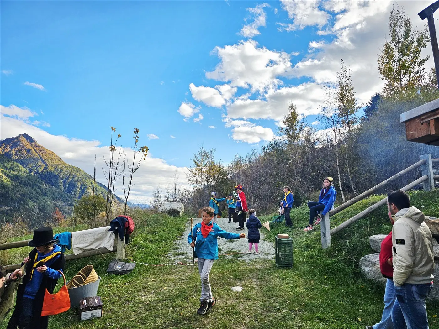 Gruppo di bambini e adulti in un'area verde all'aperto, con montagne sullo sfondo. Alcuni bambini danzano mentre altri stanno seduti o si godono il paesaggio. Cielo blu con nuvole bianche e alberi circostanti.
