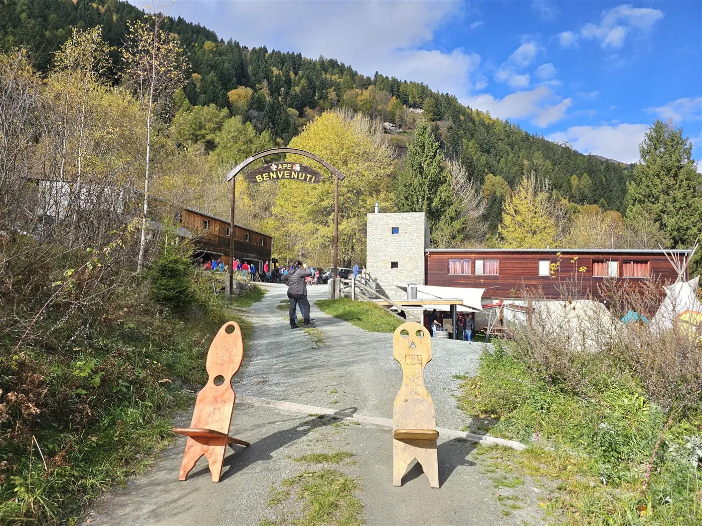 Un sentiero che conduce a un'area di accoglienza con una grande insegna "Benvenuti". Ai lati del sentiero, ci sono due grandi sedie di legno. Sullo sfondo, si vedono edifici e alberi di montagna sotto un cielo parzialmente nuvoloso.