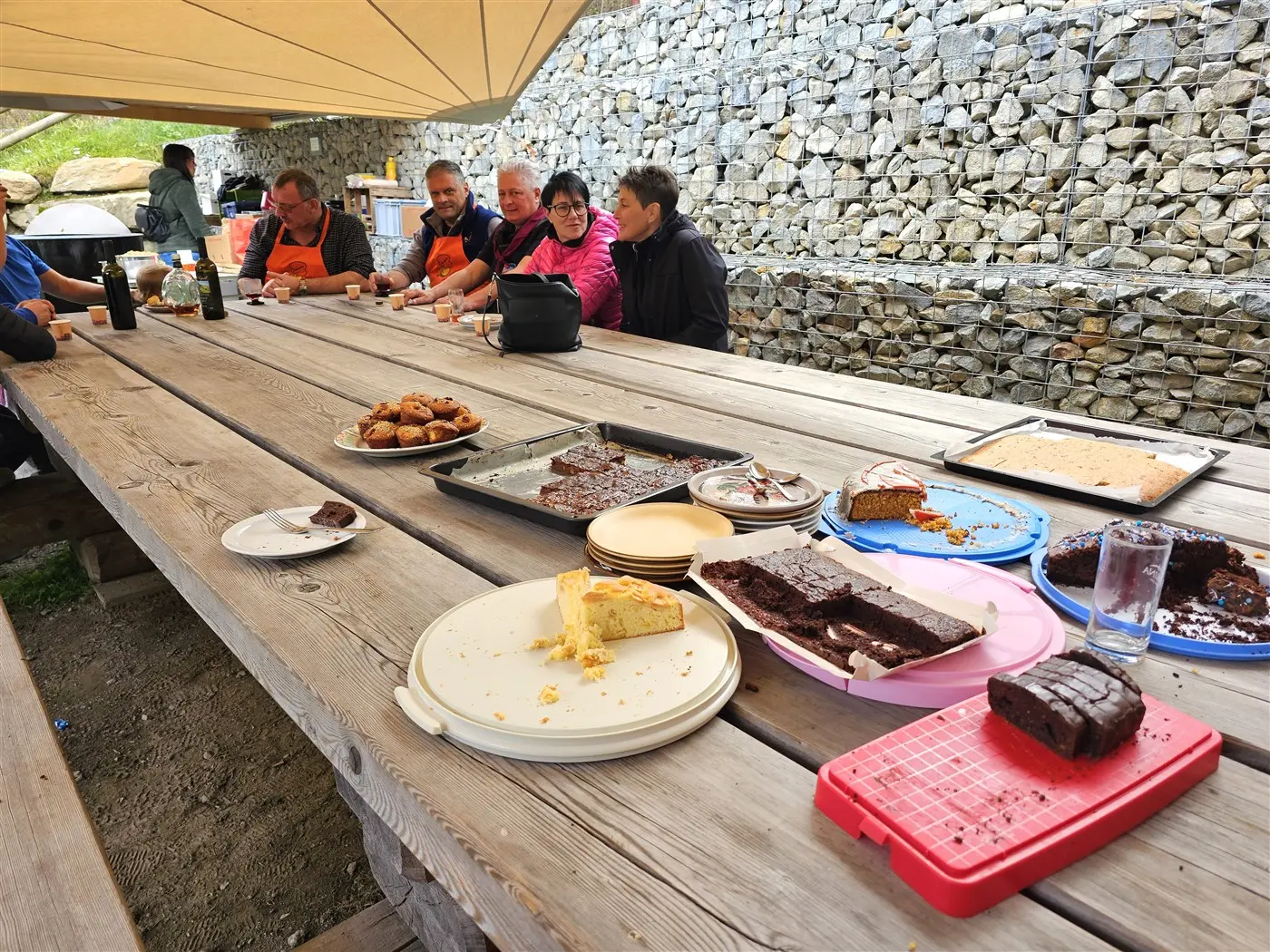 Un tavolo di legno con diversi dessert, tra cui torta al cioccolato, brownie e muffin. Sei persone sono sedute sul lato opposto del tavolo, alcune indossano grembiuli arancioni. Sullo sfondo, una parete di pietra.