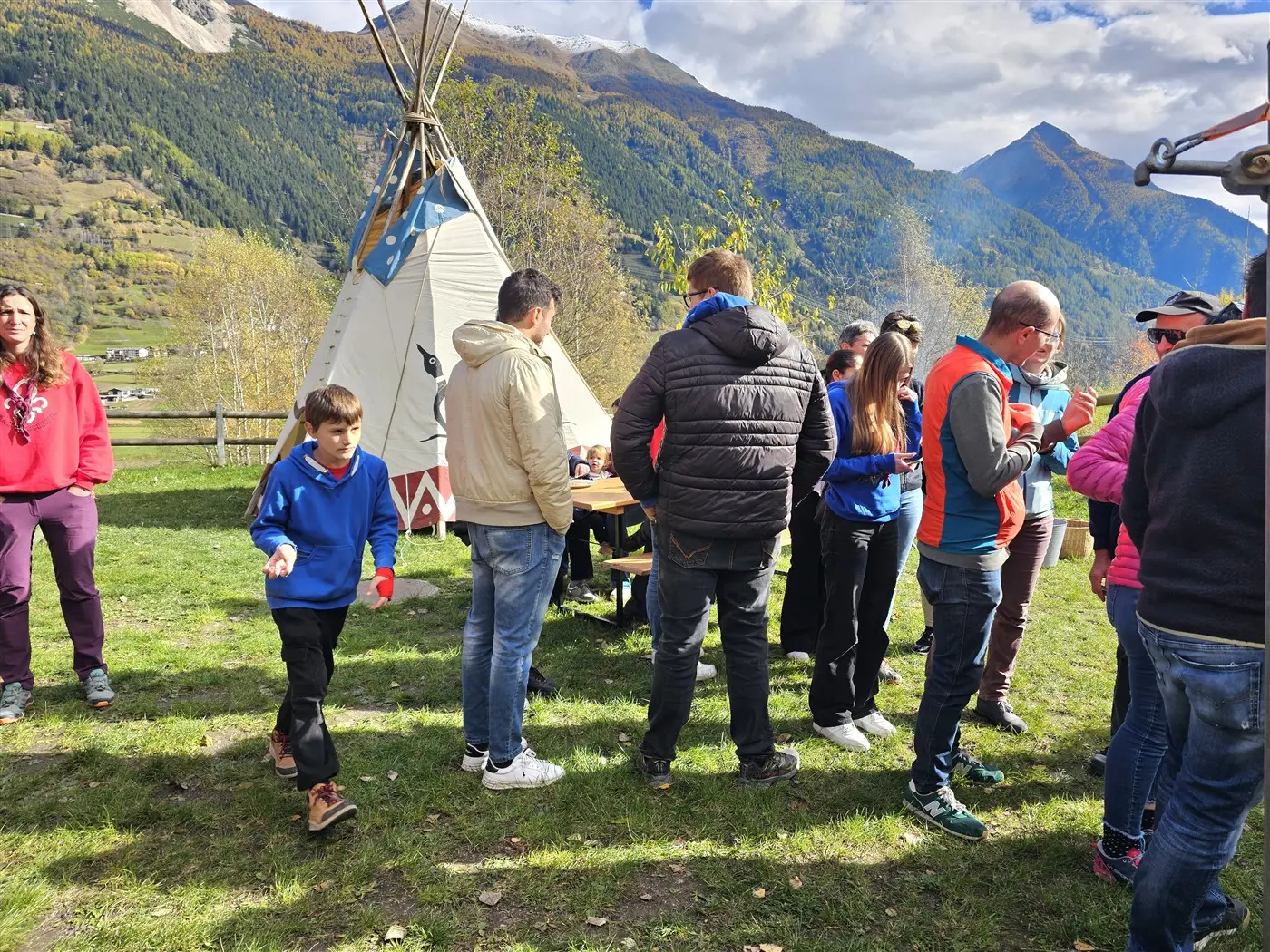 Gruppo di persone riunite all'aperto vicino a un tipì, con montagne sullo sfondo. Alcuni partecipanti indossano felpe colorate e sono impegnati in conversazioni, mentre un bambino si allontana. Il paesaggio è verde e soleggiato.