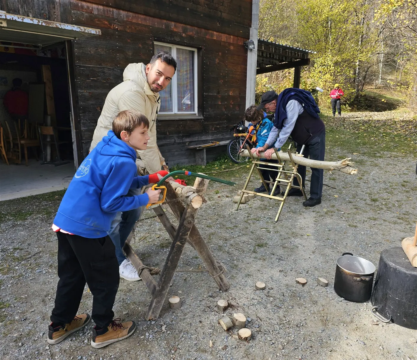 Un uomo e un ragazzo stanno segando un tronco di legno su un treppiede all'aperto, mentre altri due adulti lavorano in background. Ci sono anche alcune sedie di legno e un ambiente naturale con alberi autunnali.