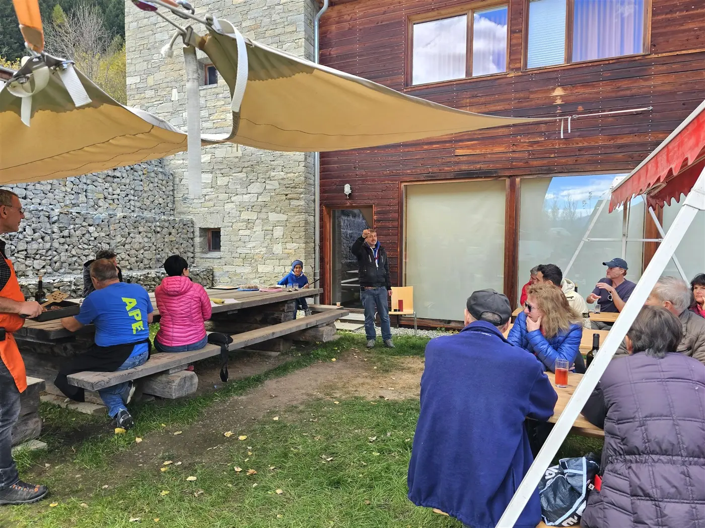 Gruppo di persone sedute a tavoli all'aperto presso una struttura in legno e pietra. Alcuni partecipanti ascoltano un uomo che parla in piedi. Ombrelloni forniscono ombra mentre il prato verde circostante completa l'ambiente.