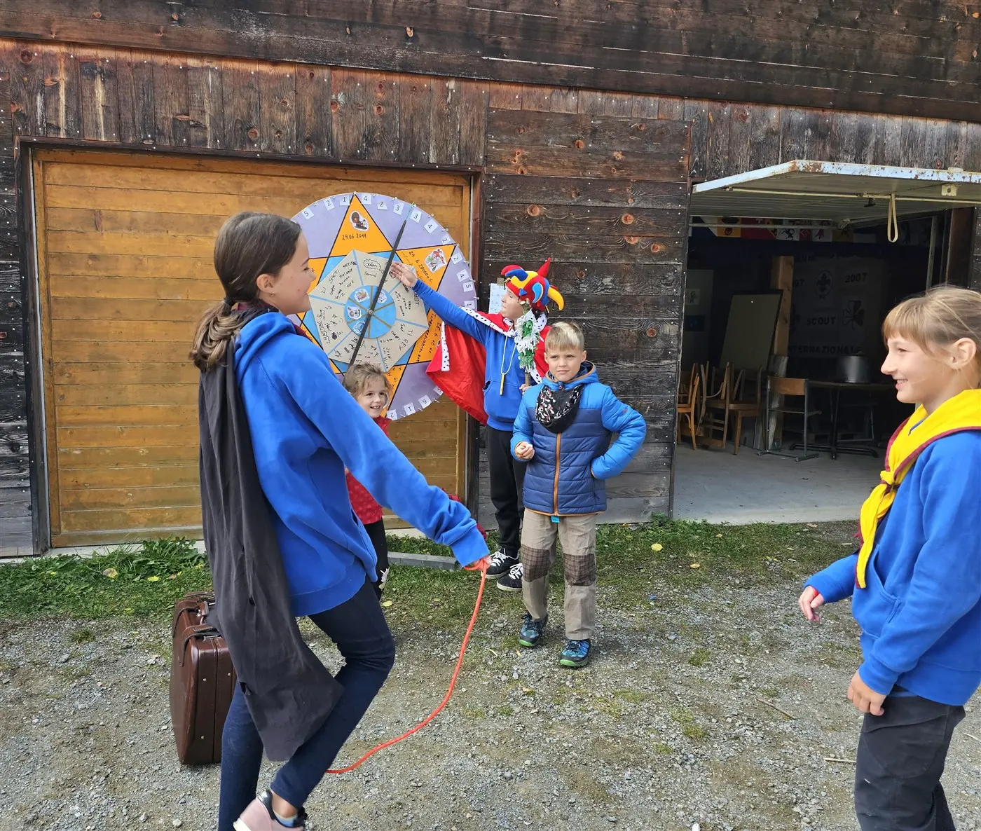 Gruppo di bambini all'aperto, alcuni indossano felpe blu. Una ragazza sta saltando la corda mentre un altro bambino gira una ruota colorata. Un terzo bambino indossa un costume creativo. Sullo sfondo si vede un edificio di legno e alcuni tavoli.