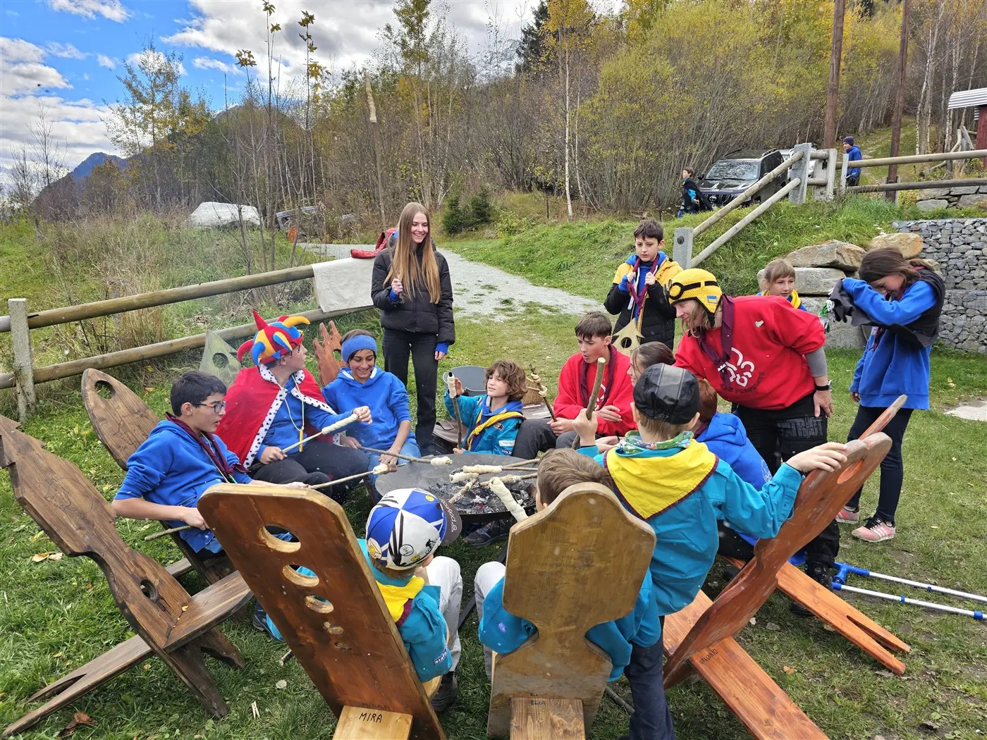 Un gruppo di bambini e adolescenti si riunisce attorno a un falò in un'area verde. Alcuni sono seduti su grandi sedie di legno, mentre altri stanno in piedi. Gli alberi circostanti e le nuvole nel cielo creano un'atmosfera di avventura.