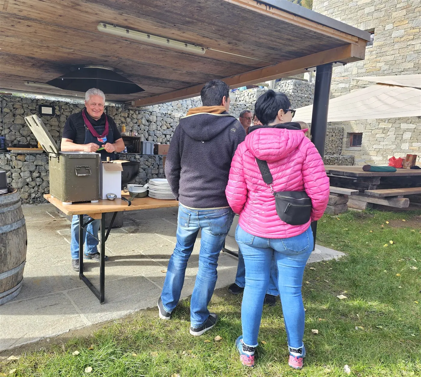Un gruppo di persone si trova sotto un pergolato all'aperto, mentre un uomo dietro un tavolo serve cibo. Due persone, di spalle, indossano giacche e jeans, mentre l'uomo mostra un sorriso e indossa un grembiule. Il contesto è caratterizzato da pietre a vista e un'atmosfera informale.