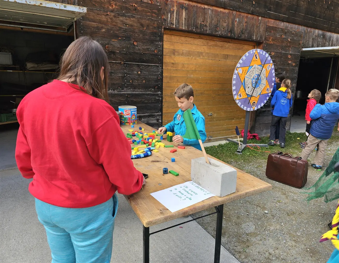 Due bambini giocano all'aperto vicino a un tavolo di legno, dove uno di loro è concentrato a impilare mattoncini colorati. Sullo sfondo, un orologio e un gruppo di altri bambini.