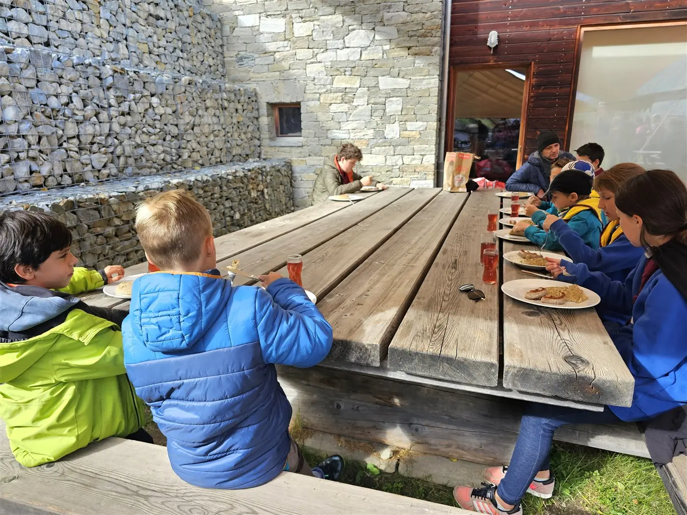 Gruppo di bambini seduti a una grande tavola di legno mentre mangiano. Alcuni sono in primo piano con piatti e bevande, altri sono più lontani e sembrano assorti. Sullo sfondo, ci sono muri in pietra e una struttura architettonica moderna.