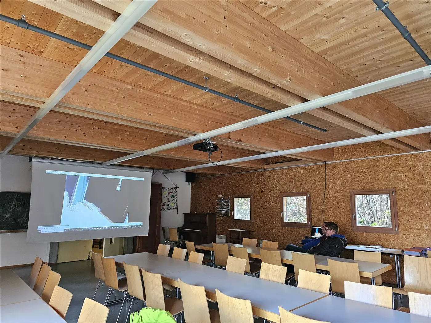 Aula spaziosa con un soffitto in legno chiaro e un grande tavolo da pranzo. Un uomo è seduto da solo vicino a un computer portatile. Sul muro è presente un proiettore che mostra un'immagine.