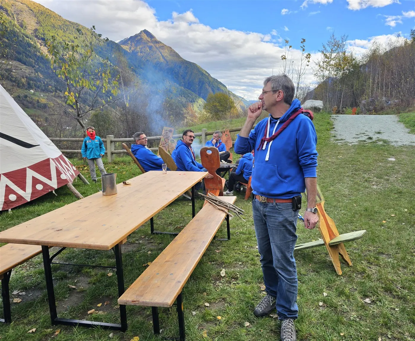 Un gruppo di persone con felpe blu si rilassa all'aperto vicino a un tavolo di legno. Sullo sfondo, ci sono monti e alberi, mentre una leggera fumata si alza da un fuoco. Una persona in primo piano sta pensando, mentre altri sono seduti e chiacchierano.
