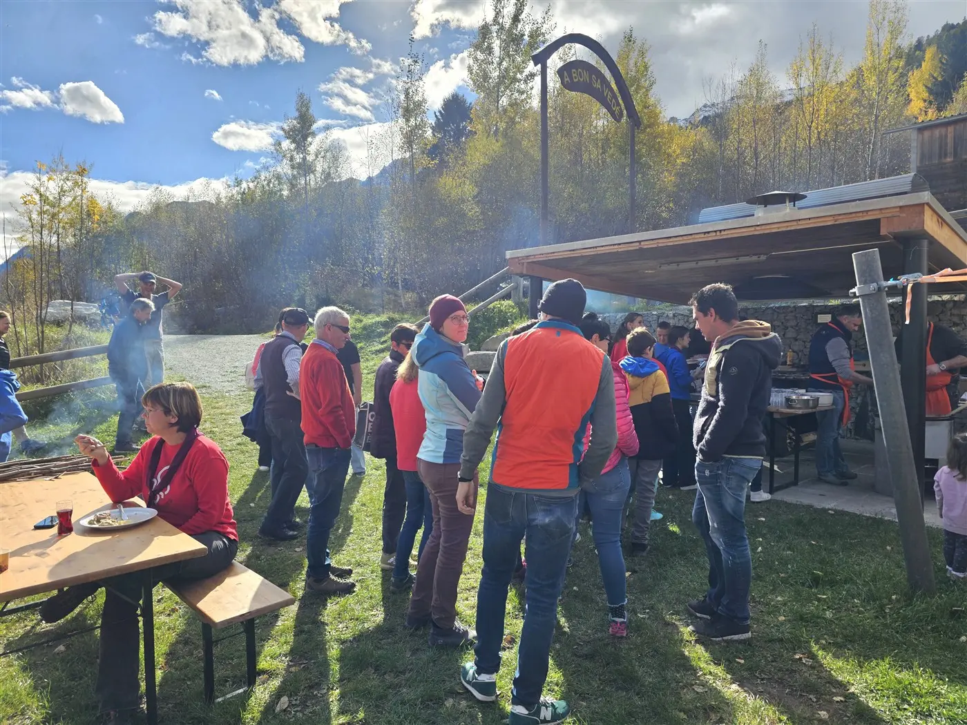 Un gruppo di persone si riunisce all'aperto davanti a un chiosco, con vari attrezzi da cucina e un'atmosfera conviviale. Alcuni partecipanti indossano giacche colorate, mentre altri chiacchierano e mangiano. Gli alberi circondano l'area, e nel cielo si vedono nuvole e raggi di sole.