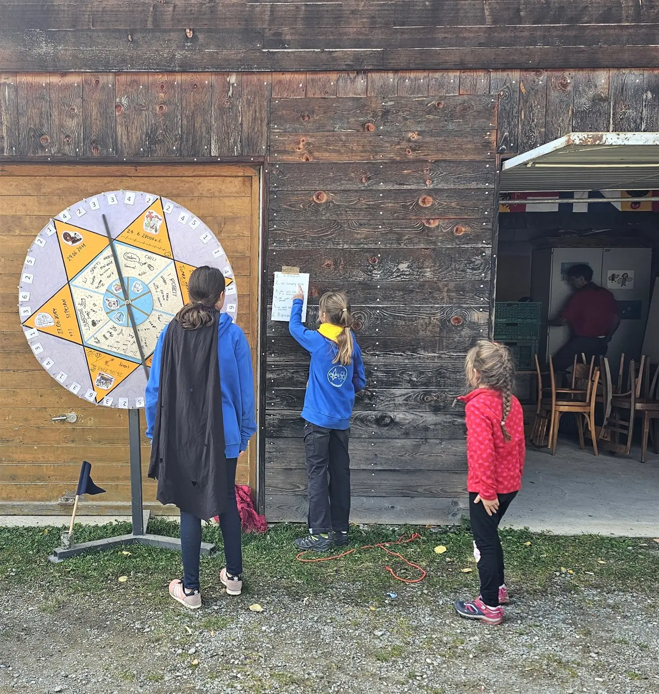 Tre bambini interagiscono con una ruota della fortuna all'aperto. Due di loro stanno leggendo un foglio attaccato al muro di legno, mentre un terzo bambino è di spalle. Si intravede una stanza con sedie all'interno.