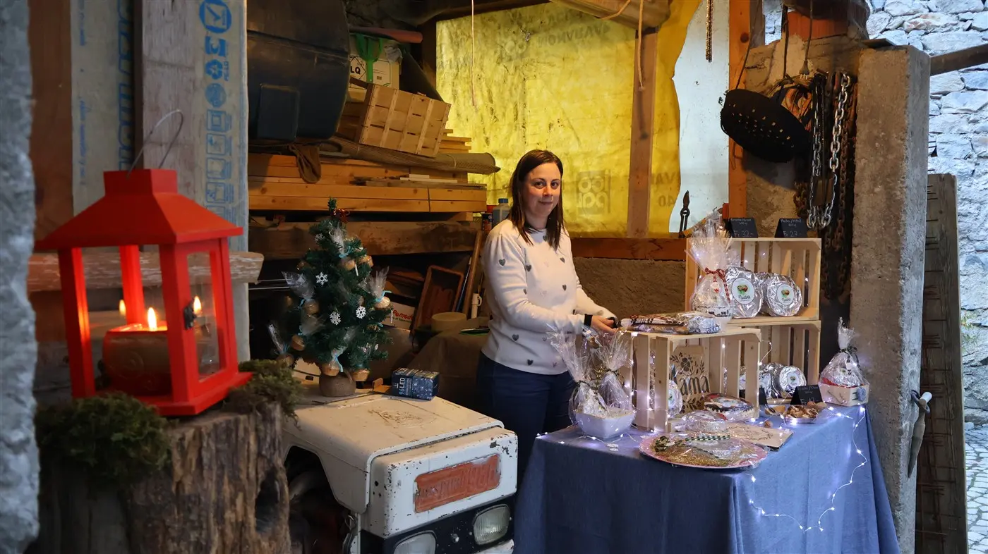 Una donna sorride mentre sistema prodotti artigianali su un tavolo in un piccolo negozio rustico. Sullo sfondo si intravedono decorazioni natalizie, tra cui un al