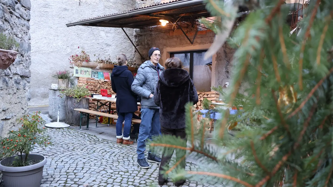 Un cortile acciottolato con persone che conversano vicino a tavoli all'aperto. Una persona indossa un giubbotto grigio e occhiali. Sullo