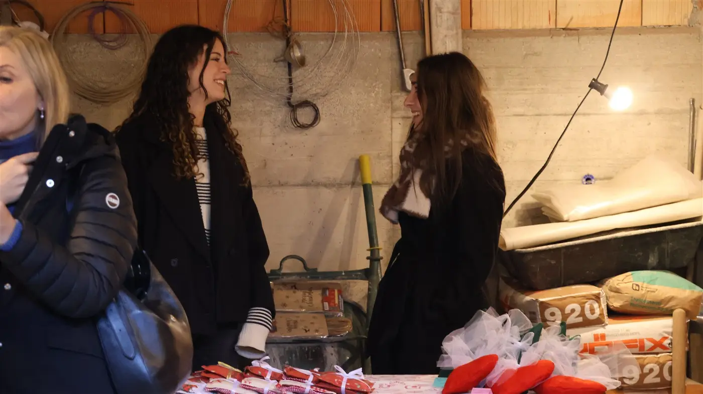 Due donne sorridono e conversano in un mercatino all'interno di un edificio. Sul tavolo davanti a loro ci sono decorazioni natalizie. Altre persone sono visibili sullo