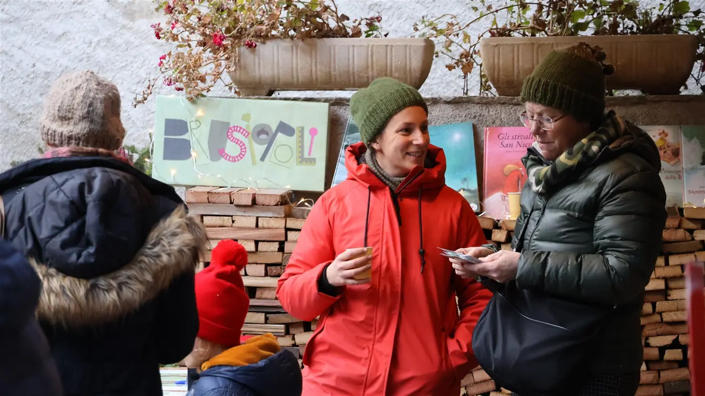 Due donne conversano in una fiera, una indossa una giacca rossa e tiene una bevanda, mentre l'altra controlla il telefono. Sullo sfondo si vedono cartelli