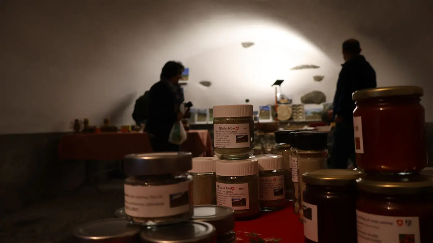 Mercato in un ambiente interno con tavoli espositivi. Barattoli di conserve e spezie in primo piano, mentre due persone stanno osservando i prodotti sullo sfondo. luce soff