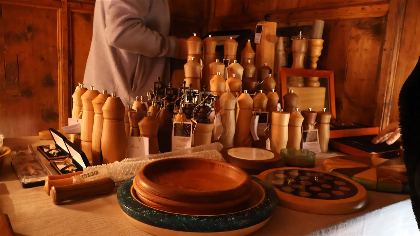 Una esposizione di oggetti in legno, tra cui macina pepe e ciotole, sistemati su un tavolo. Sullo sfondo, una persona sta selezionando gli og