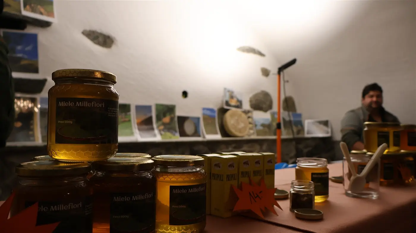 Vasi di miele millefiori esposti su un tavolo con fotografie e oggetti sullo sfondo. Un uomo è visibile in lontananza.