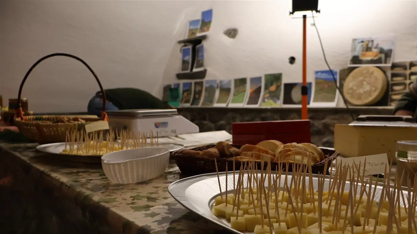 Tavolo imbandito con vari formaggi a cubetti e altri snack, circondato da foto appese su una parete. Una cesta di vimini contiene prodotti assortiti e ci