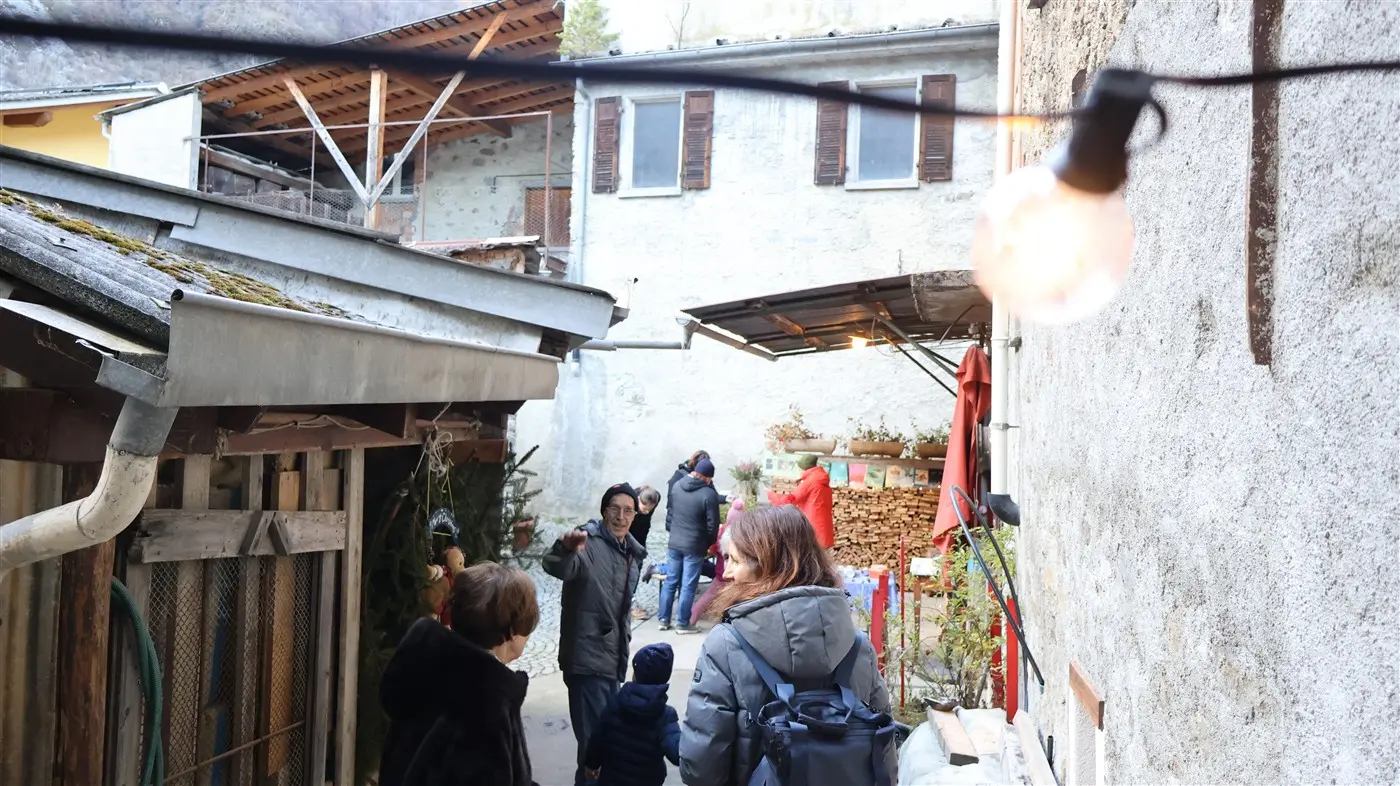 Un cortile affollato con persone che passeggiano e chiacchierano. Edifici di pietra sullo sfondo e luci decorative appese sopra. Si vedono