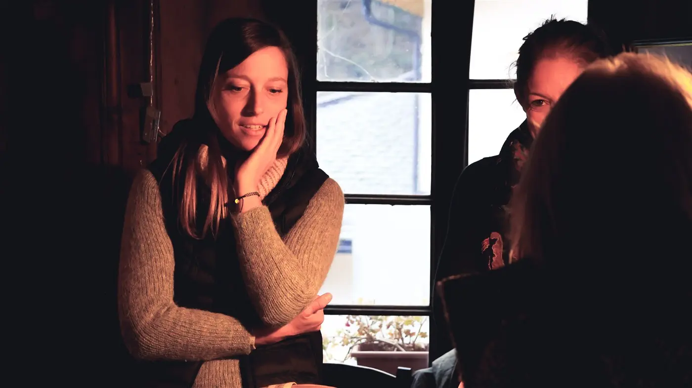 Due donne sedute in un ambiente accogliente. Una ha le braccia incrociate e sembra pensierosa, mentre l'altra la osserva con un sorriso. La luce tenue crea