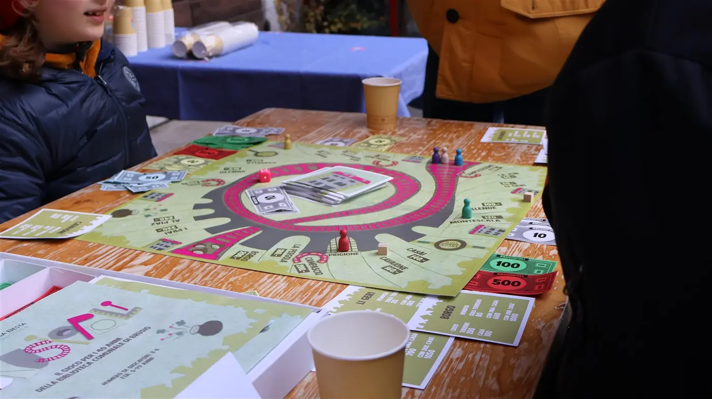 Un tavolo di legno con un gioco da tavolo in corso. Il gioco presenta una grande mappa circolare con vari elementi come case, banconote di diverse denominazioni e segnal