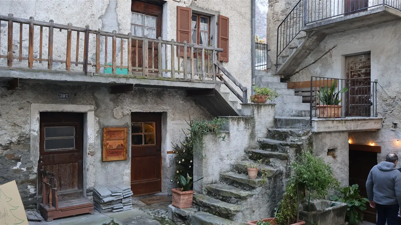 Un cortile di un antico edificio in pietra, con scale che conducono a diversi piani. Porte in legno e finestre con persiane si affacciano sulla scena. Vasi