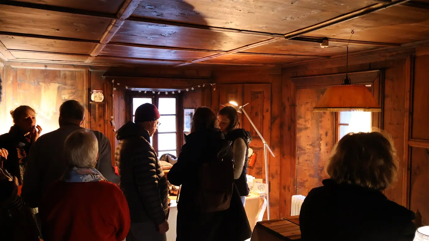 Un gruppo di persone in una stanza con pareti in legno, mentre conversano. Sono presenti una lampada a sospensione e una luce che filtra dalle finestre.