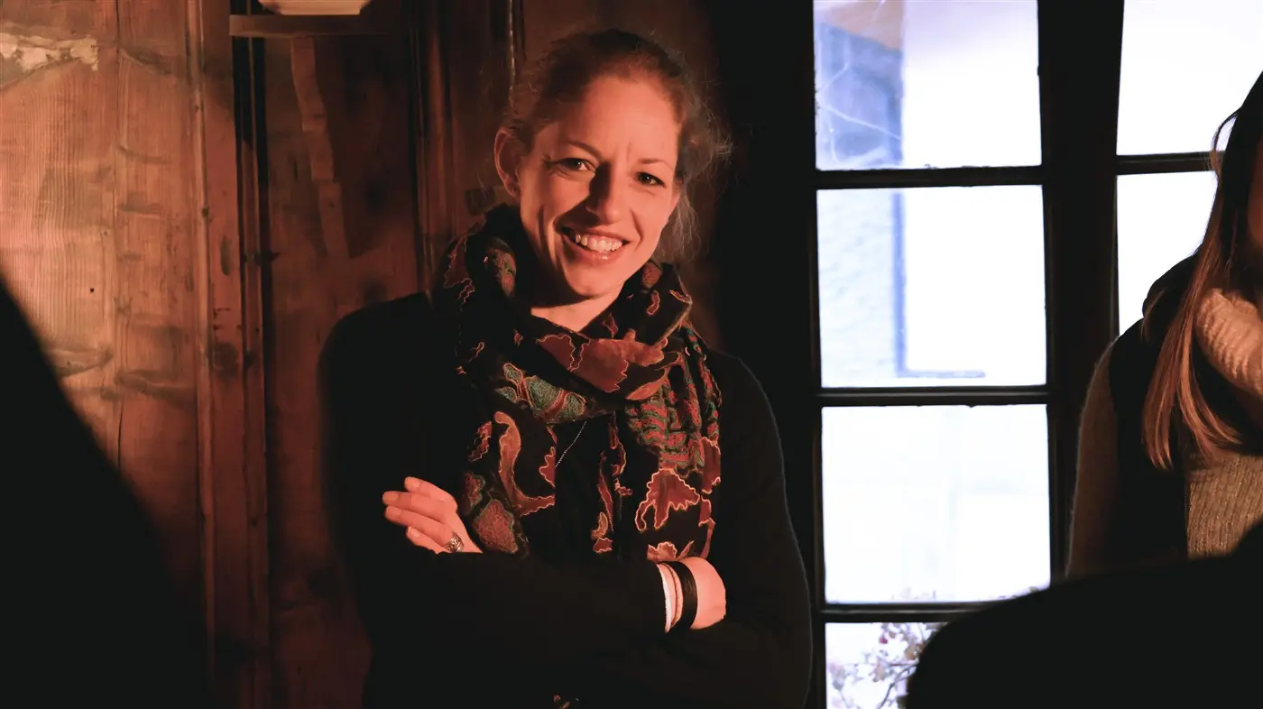 Una donna sorridente con una sciarpa colorata, mentre è appoggiata a una parete di legno scuro. Alle sue spalle ci sono finestre con luce naturale che entra