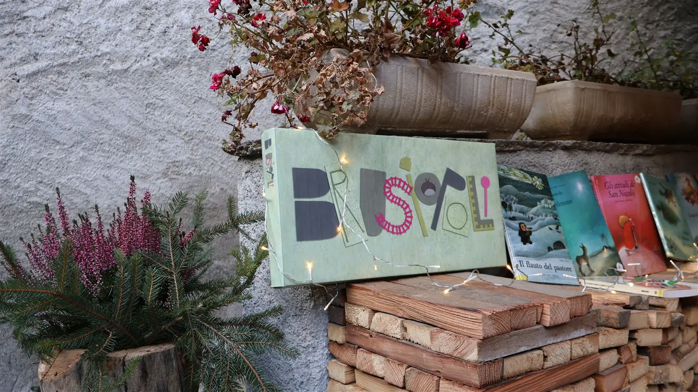 Un libro intitolato "Bruscoli" è poggiato su un impilamento di legno, circondato da piante ornamentali. Luci decorative brillano attorno al