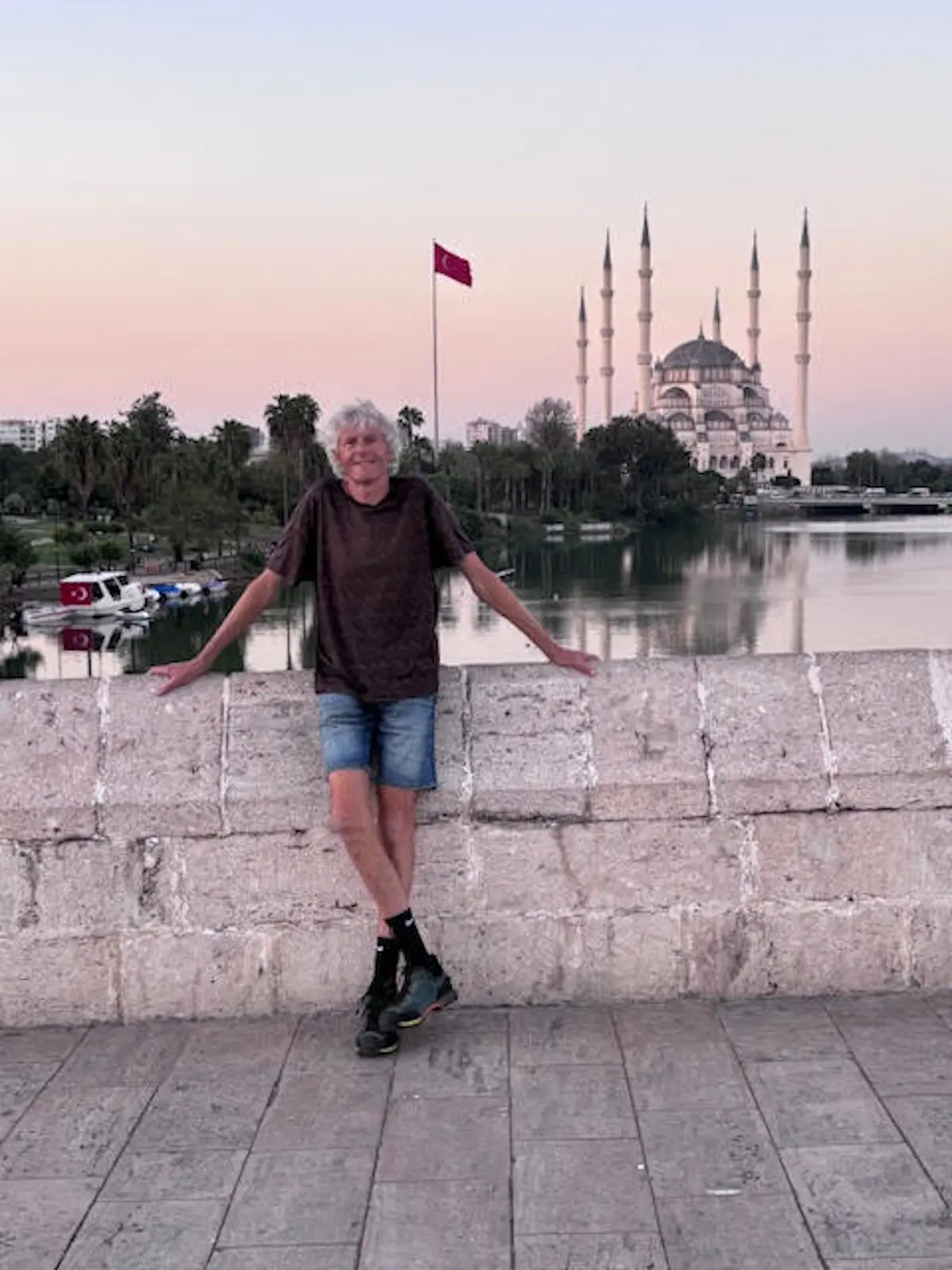 Una persona sorridente in piedi su una barriera con il braccio appoggiato. Sullo sfondo, una grande moschea con minareti e una bandiera turca che sventola. È il tramonto e il cielo ha toni rosa e arancioni.