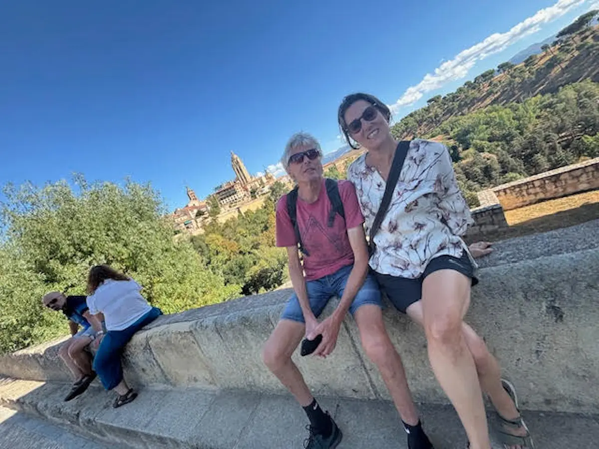 Due persone sedute su un muraglione, con uno sfondo panoramico di una città collinare. Il cielo è sereno e il paesaggio è verde e collinare.