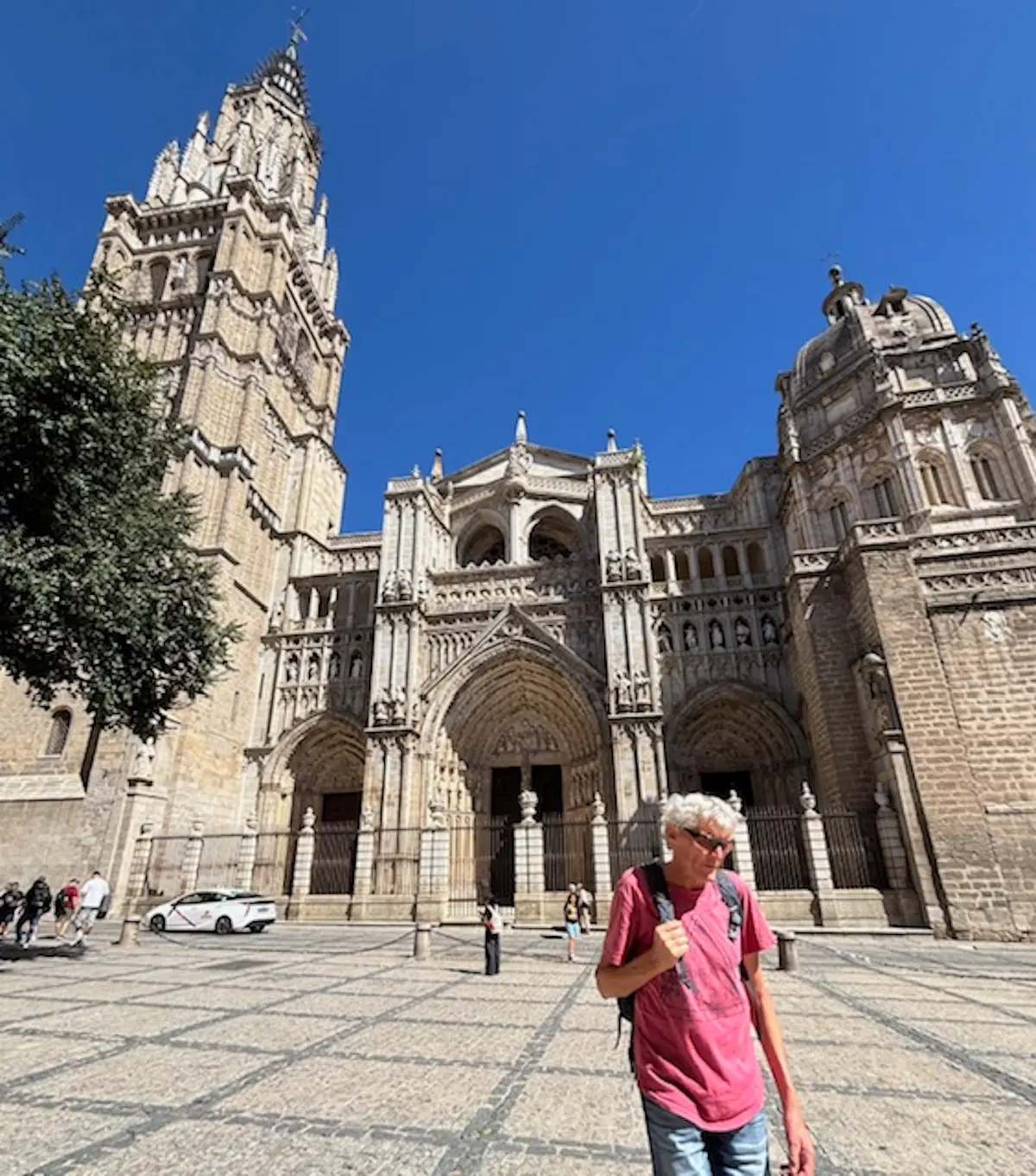 Una persona con capelli grigi cammina davanti a una cattedrale imponente con torri e un cielo blu chiaro. Alcuni turisti sono visibili intorno.